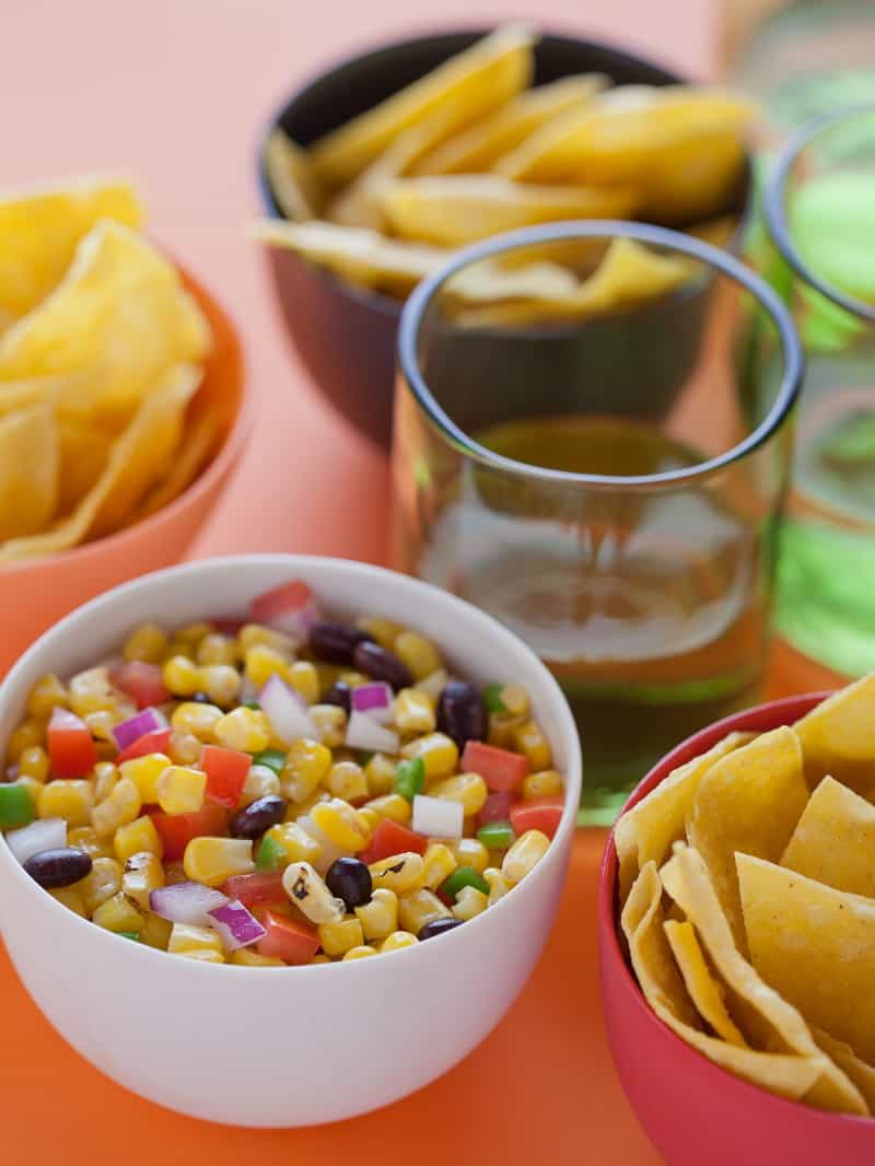 A close up of a bowl of grilled corn salsa next to bowls of tortilla chips.