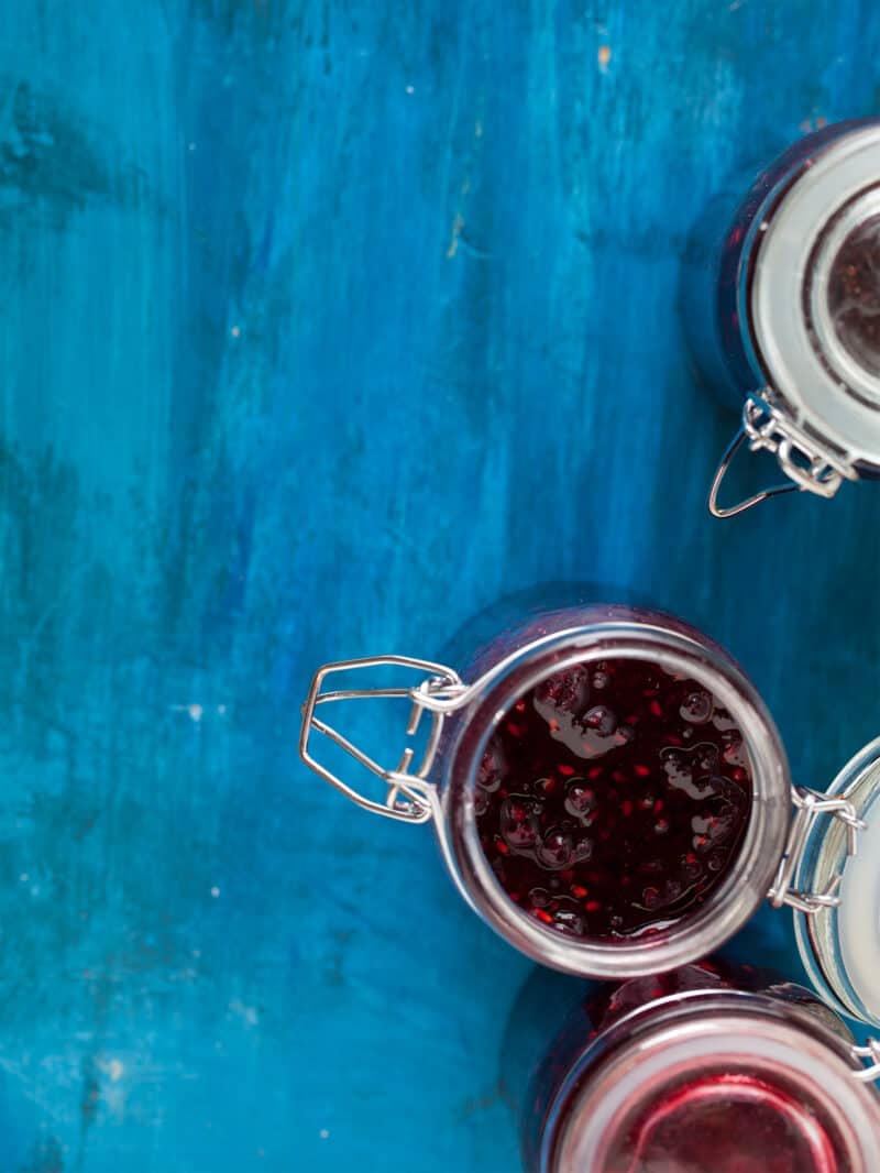 An open glass jar next to closed jars of simple blackberry jam on a blue surface.