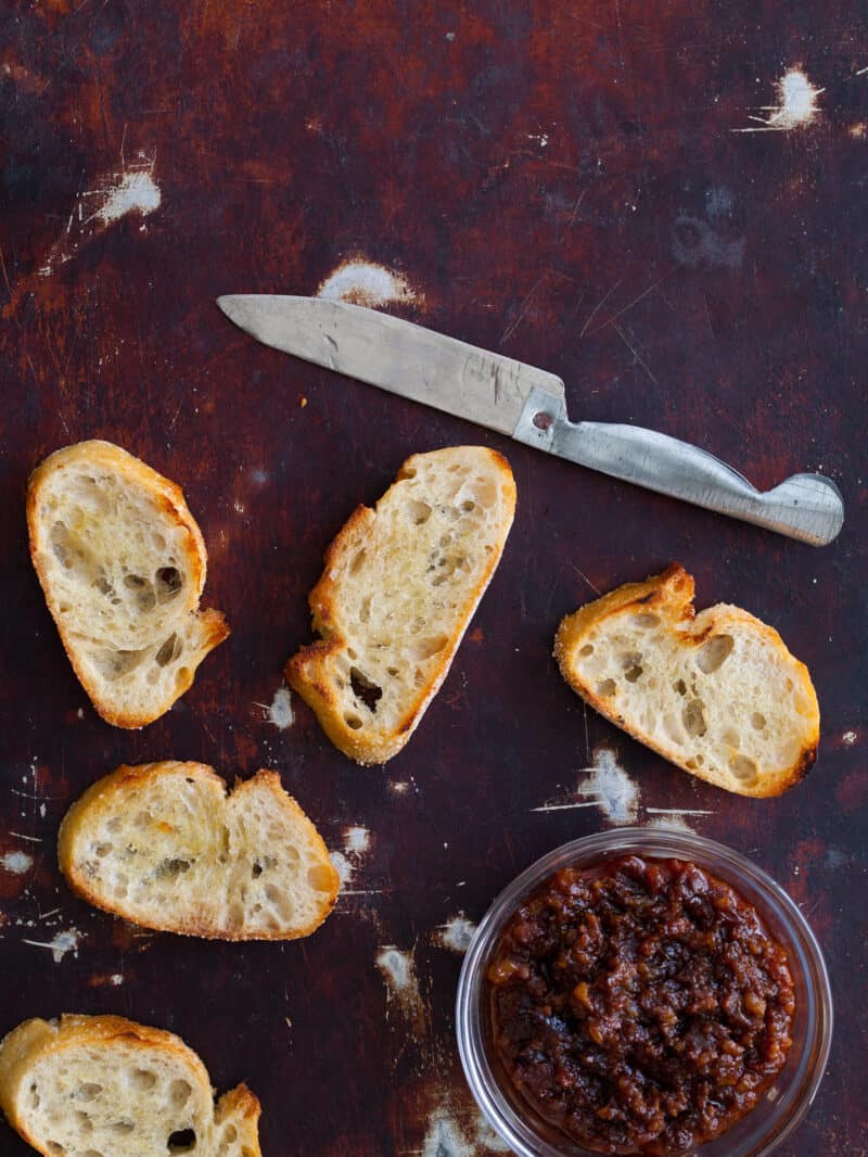 Crostini with a knife and a bowl of boozy bacon jam.