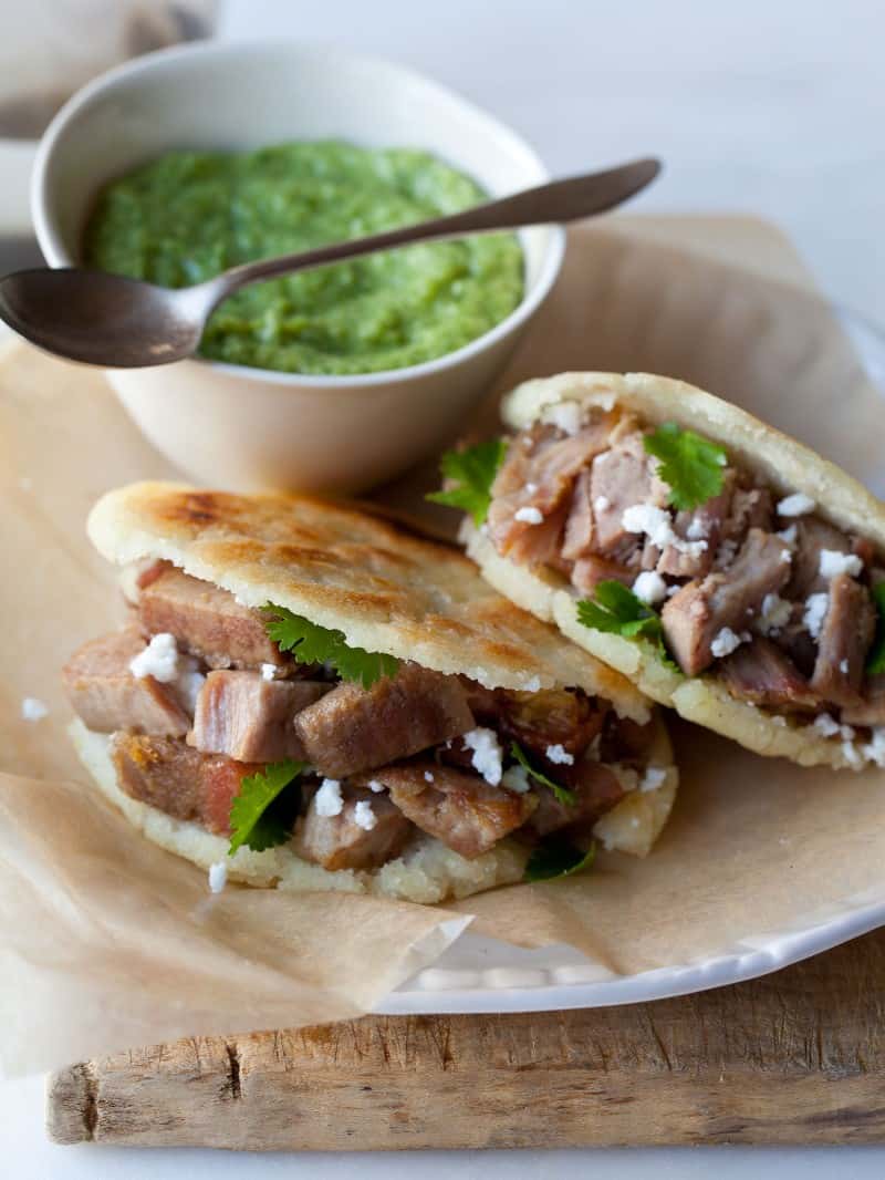 A plate of arepas filled with carnitas and a bowl of guasacaca with a spoon.