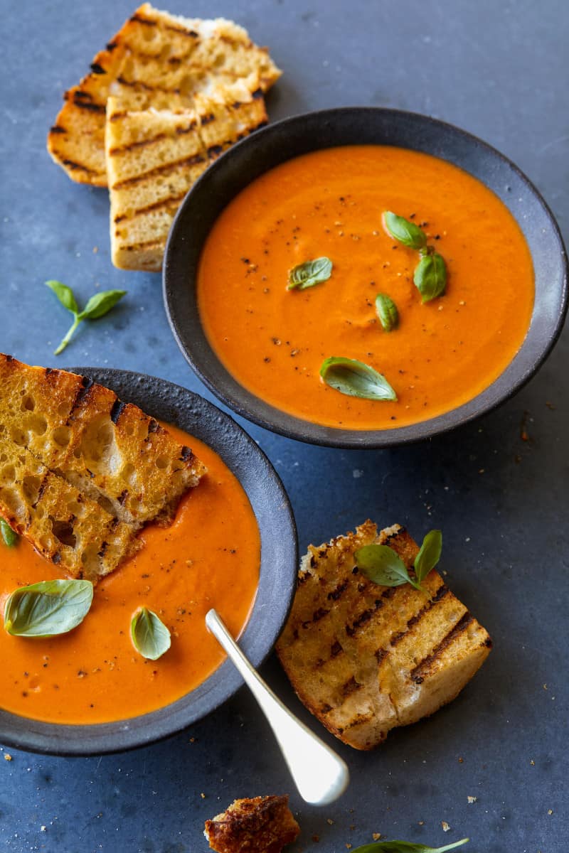 Bowls of creamy roasted tomato and basil soup with grilled bread and fresh basil.