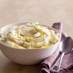 A bowl of mashed potatoes with butter and chives, a purple napkin, and a spoon.