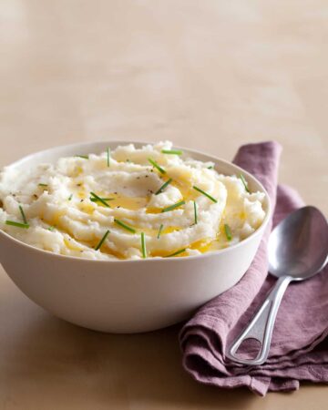 A bowl of mashed potatoes with butter and chives, a purple napkin, and a spoon.