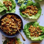 A bowl of chicken with a spoon next to lettuce wraps on small plates.