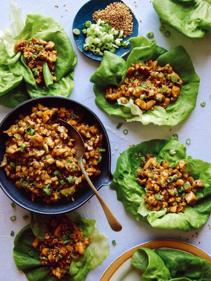 A bowl of chicken with a spoon next to lettuce wraps on small plates.