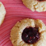 A close up of peanut butter and jelly potato chip thumbprint cookies on a pink surface.