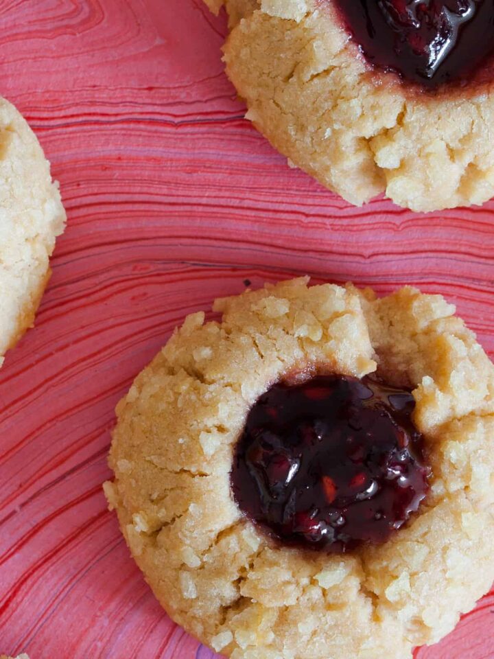 A close up of peanut butter and jelly potato chip thumbprint cookies on a pink surface.