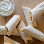 Quick and easy baked chicken flautas with a ramekin of sour cream on a wooden surface.