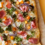 A close up of prosciutto kale butternut squash pizza on a wooden cutting board.