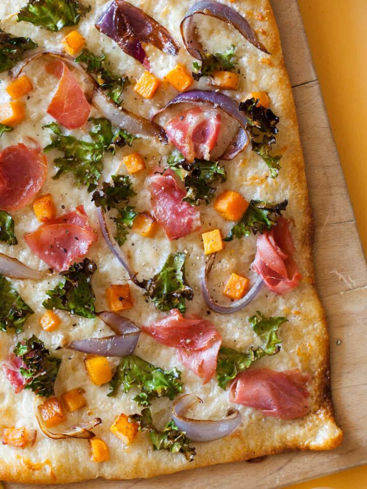 A close up of prosciutto kale butternut squash pizza on a wooden cutting board.