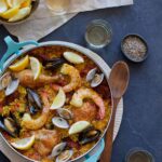 A pot of paella with spices in bowls, lemon wedges, and a wooden spoon.