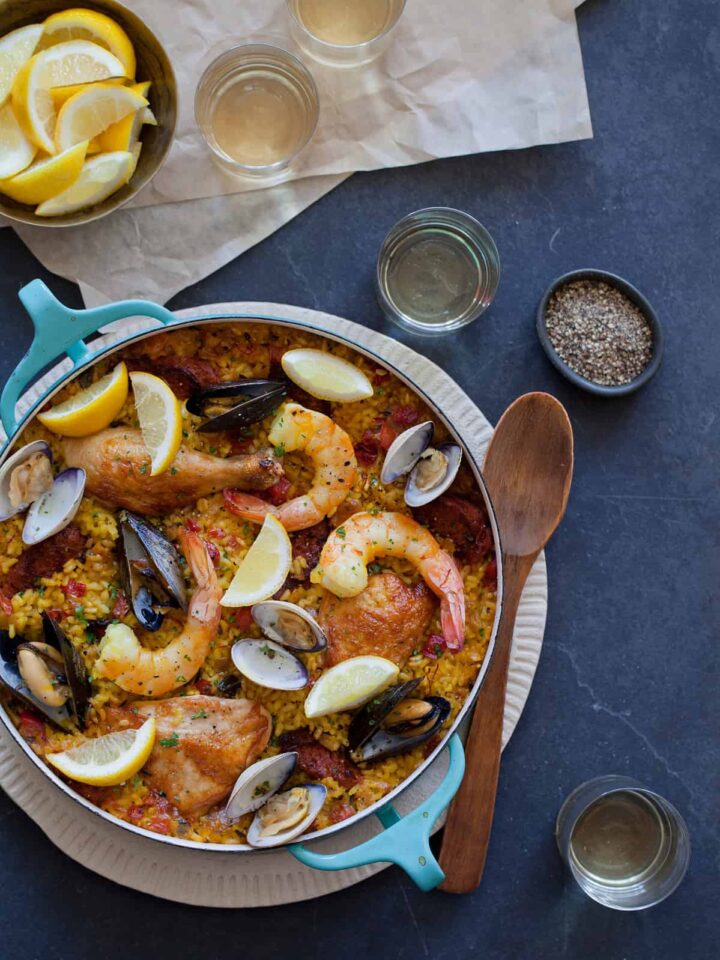 A pot of paella with spices in bowls, lemon wedges, and a wooden spoon.