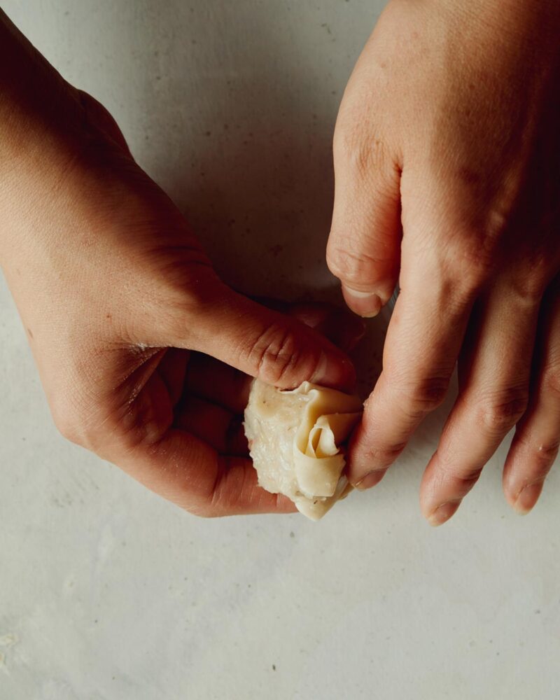 Step 5 of making shrimp shumai. Gently wet your fingers in your bowl of water. Stick the edges of the wonton wrapper that are resting on top of your hand down to the sides on the shrimp shumai. You may need to re-dip your fingers. The water works as an adhesive between the layers of wonton.