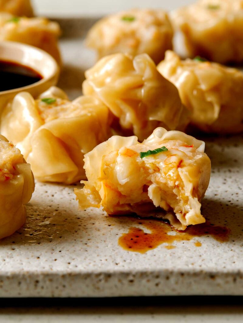 9 steamed shrimp shumai next to a ramekin of sauce. 1 has been dipped in sauce with a bite out of it