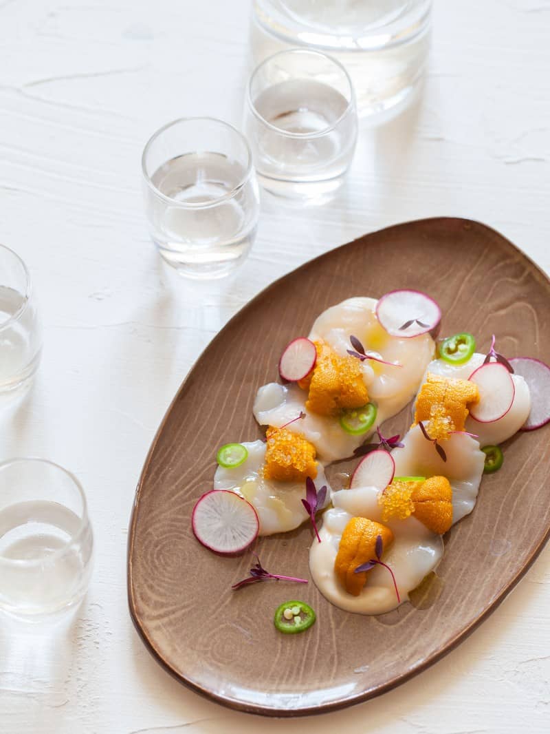 A platter of scallop and uni crudo surrounded by several drinks.