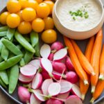 A close up of a bowl of roasted onion dip on a platter of crudités.