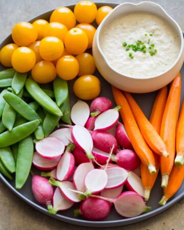 A close up of a bowl of roasted onion dip on a platter of crudités.