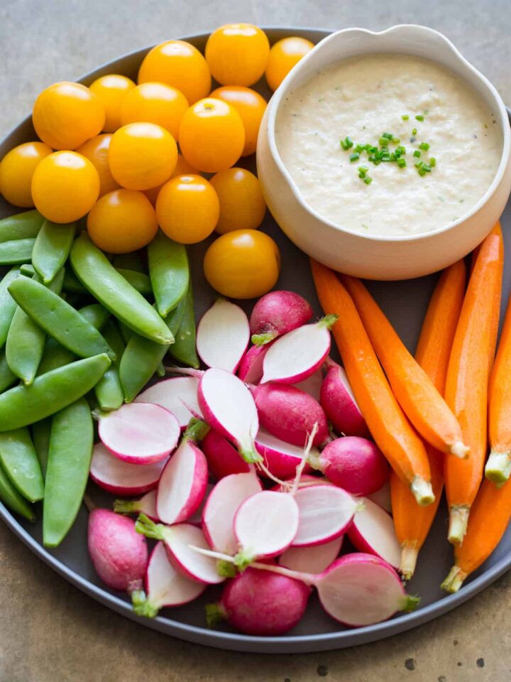 A close up of a bowl of roasted onion dip on a platter of crudités.