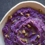 A close up of mashed purple sweet potatoes with toasted pecans in a bowl.