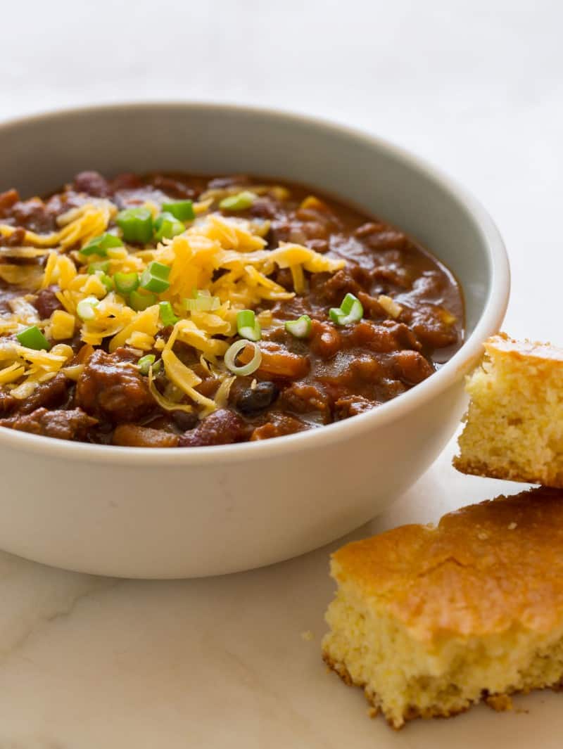 A bowl of kitchen sink chili topped with cheese and chives, with two pieces of corn bread next to it.