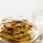 A stack of chocolate chip, bacon, orange zest pancakes drizzled in syrup with a fork.