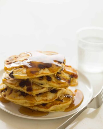 A stack of chocolate chip, bacon, orange zest pancakes drizzled in syrup with a fork.