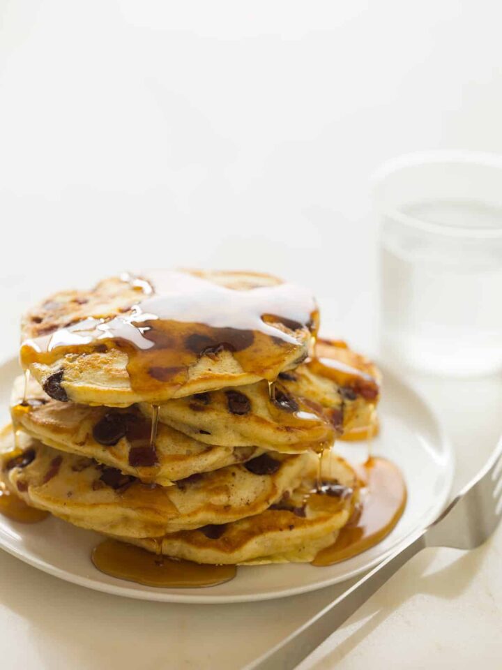 A stack of chocolate chip, bacon, orange zest pancakes drizzled in syrup with a fork.