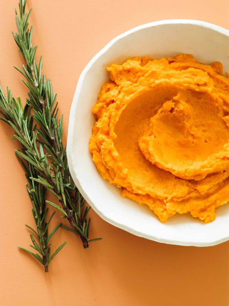 A close up of a bowl of mashed sweet potato next to a sprig of rosemary.