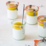 Several glass jars of vanilla bean panna cotta with mango coulis with a spoon.