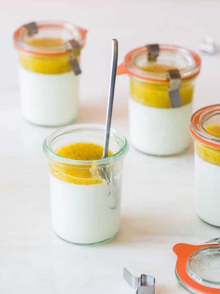 Several glass jars of vanilla bean panna cotta with mango coulis with a spoon.