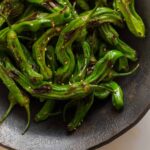 A close up of blistered shishito peppers in a dark bowl.