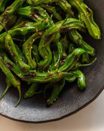 A close up of blistered shishito peppers in a dark bowl.