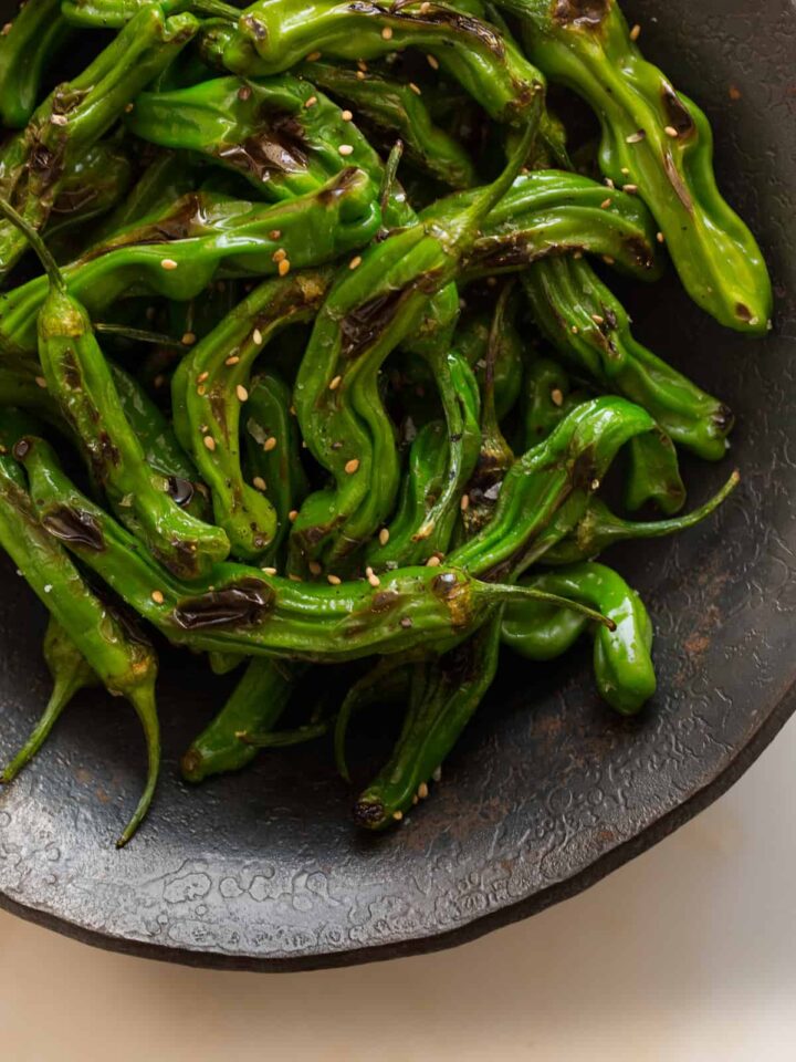 A close up of blistered shishito peppers in a dark bowl.