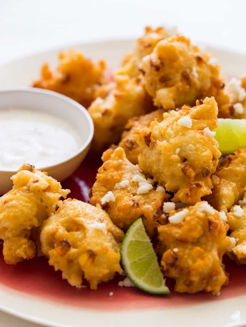 A close up of elote fritters with lime wedges and a ramekin of cilantro mayo.