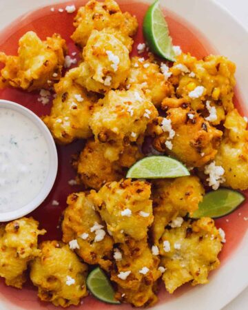 A close up of elote fritters with lime wedges and a ramekin of cilantro mayo.
