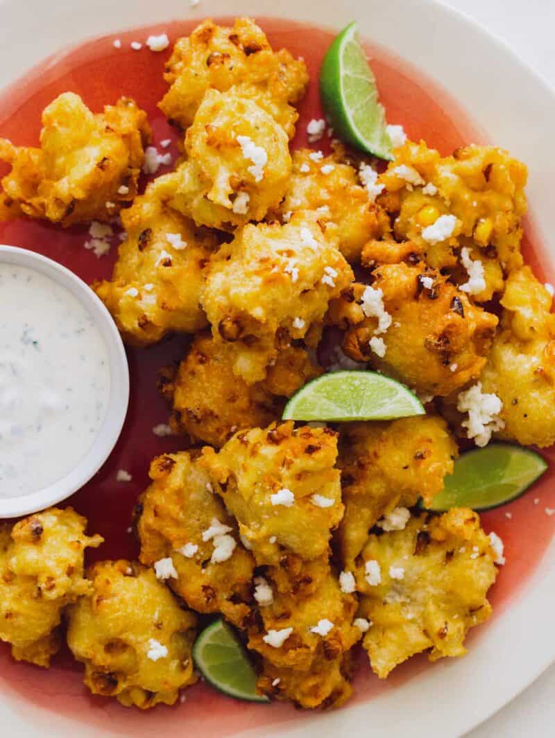 A close up of elote fritters with lime wedges and a ramekin of cilantro mayo.