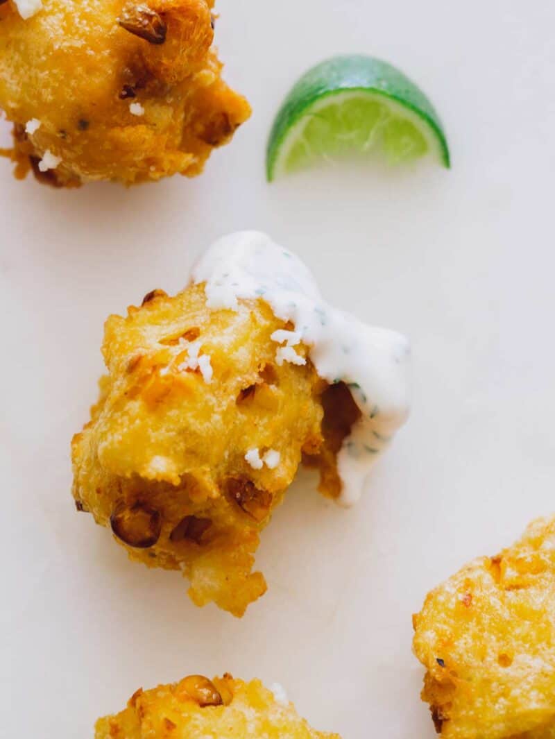 A close up of elote fritters dipped in cilantro mayo with lime wedges.