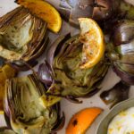 A close up of grilled artichokes with orange and lemon wedges and aloe on the side.