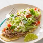 Huaraches with carnitas and fixings on a plate with a lime wedge and a fork.