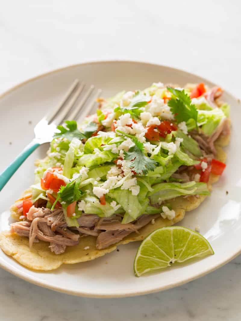 Huaraches with carnitas and fixings on a plate with a lime wedge and a fork.