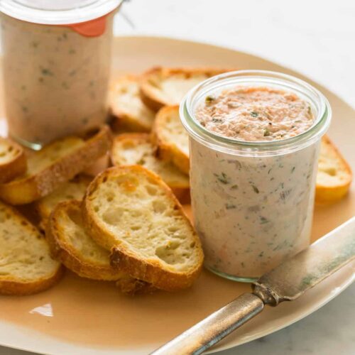 Glass jars of salmon rillettes on a plate with crostini and a knife.