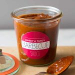 A labeled glass jar of sweet and tangy BBQ sauce, the lid, and a coated spoon.