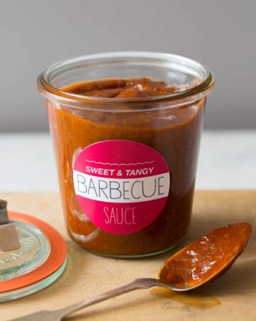 A labeled glass jar of sweet and tangy BBQ sauce, the lid, and a coated spoon.