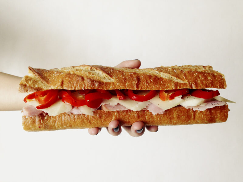 A whole bocadillo of ham, cheese, and pickled cherry peppers on a baguette in a hand.