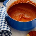 A pot of marinara sauce next to a coated wooden spoon and blue gingham linen.