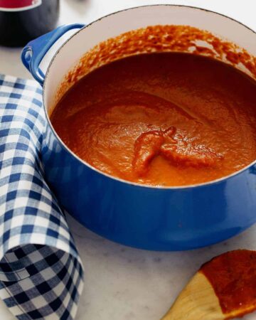 A pot of marinara sauce next to a coated wooden spoon and blue gingham linen.