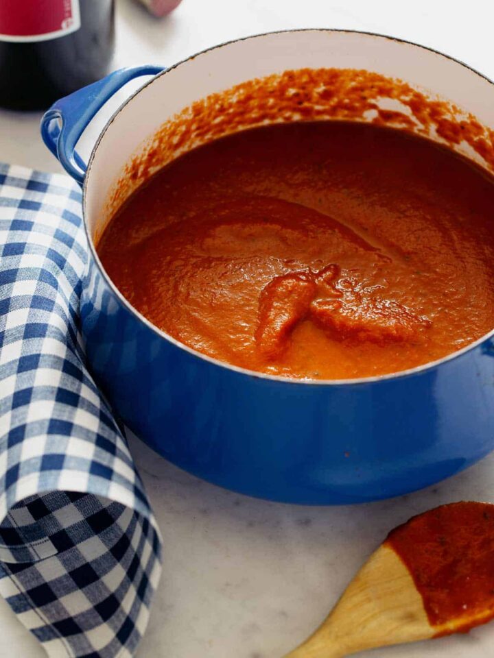 A pot of marinara sauce next to a coated wooden spoon and blue gingham linen.
