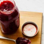 An open jar of black plum preserve next to a spoonful on a wooden cutting board.