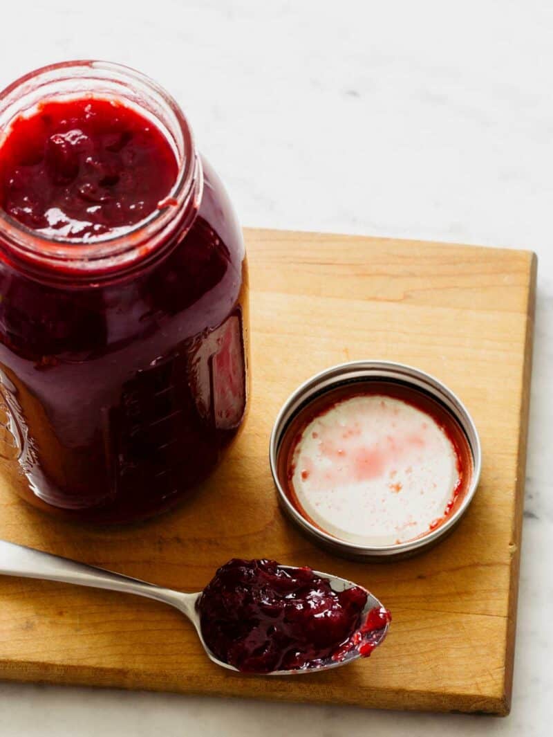 An open jar of black plum preserve next to a spoonful on a wooden cutting board.