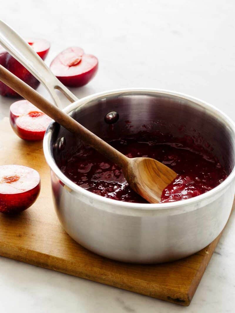 A pan of black plum preserve with a wooden spoon and fresh halved black plums.